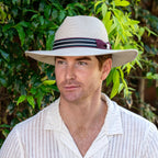Man wearing a beige fedora with a dark band and purple flower, standing against a green leafy background.