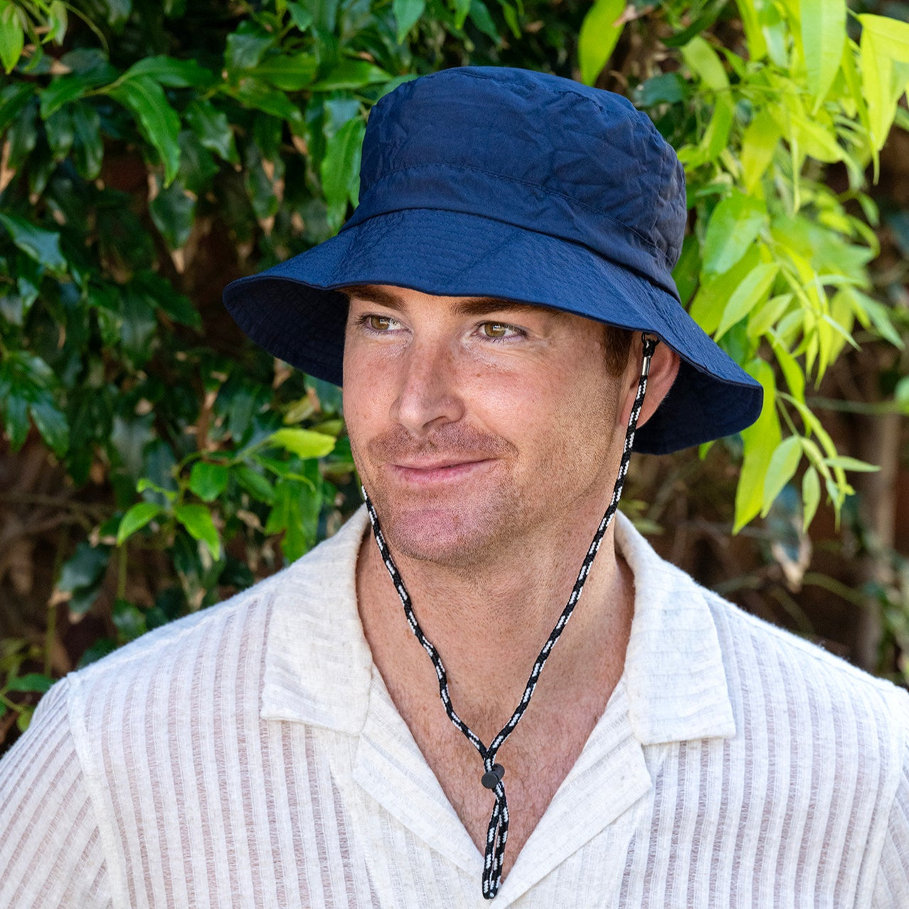 Man wearing a navy bucket hat and white shirt with green foliage in the background