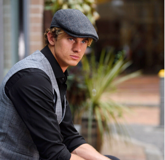 A man wearing a gray flat cap, a gray plaid vest, and a black long-sleeve shirt, looking off-camera in an outdoor urban setting.