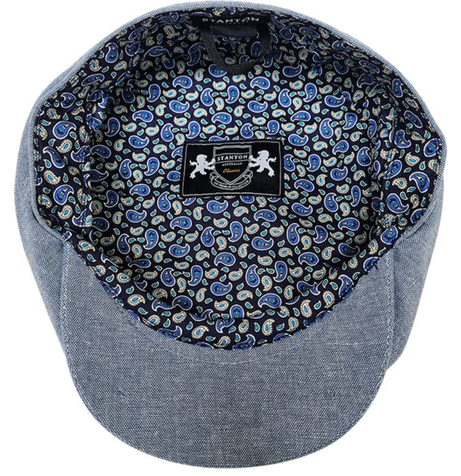 A close-up of a newsboy cap's interior and brim, showing a blue and white paisley pattern lining with a central black brand patch reading Stanton. The brim and side panels are made of a textured blue fabric.