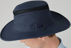 A close-up side view of a person wearing a navy blue wide-brim sun hat with a black mesh band and a small black Tilley logo tag on the side, against a light gray background.