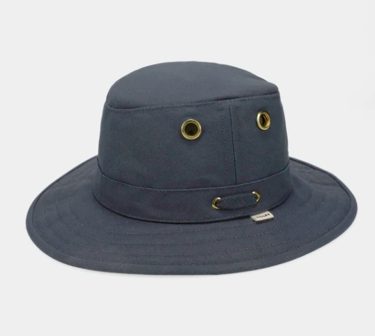 A dark slate blue bucket hat with brass grommets and a small Tilley logo tag on the side, shown against a light gray background.